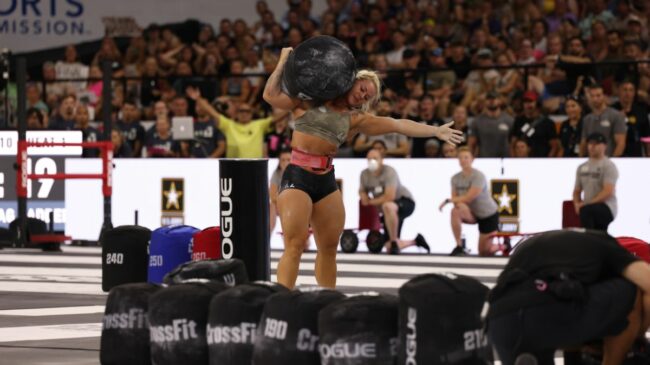 CrossFit athlete Dani Speegle, wearing blck shorts and camo tank top, holds a heavy sandbag on her shoulder.