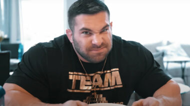 A scowling bodybuilder wearing a T-shirt and eating his lunch.