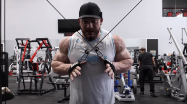 A bodybuilder in a sleeveless shirt wearing a hat. He's working out his triceps.