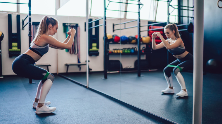 A person looks into a mirror while performing a bodyweight mini band squat.