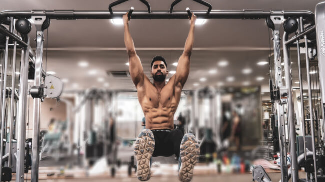 A shirtless person performs hanging leg raises in the gym.