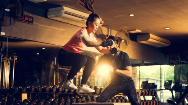 A person with a ponytail performs a box jump while their trainer looks on.