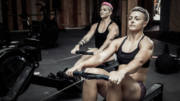 Two people with short hair wear a sports bra and tank top as they row on rowing machines.