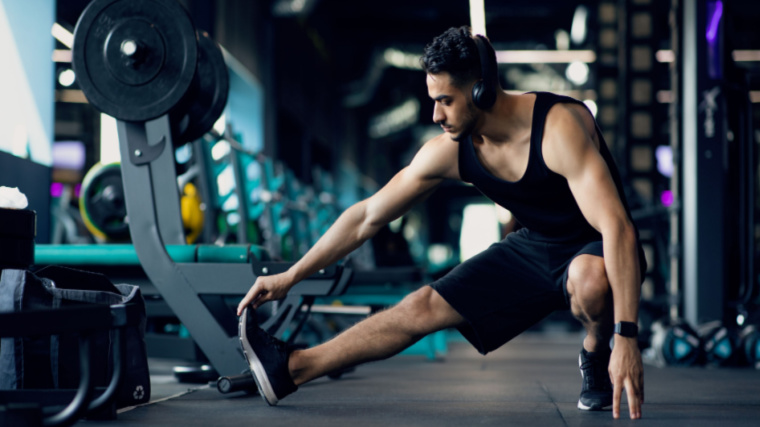 A person wears a black tank top, black shorts, and black headphones while stretching their leg leg in a lateral lunge.