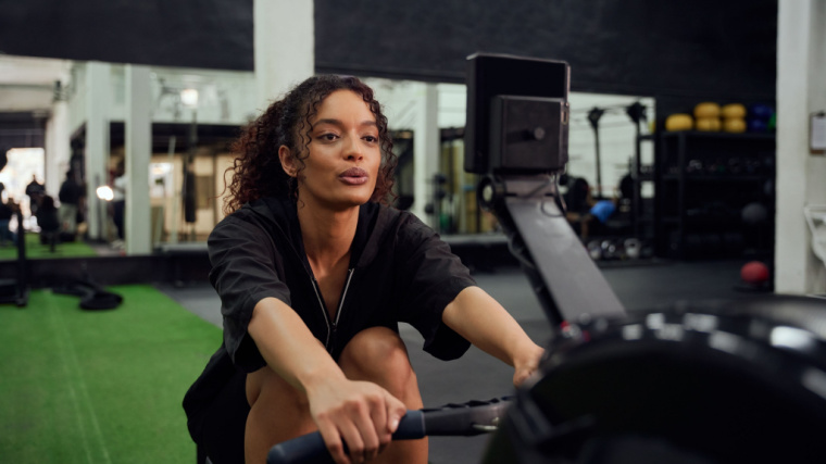 A person wears a black t-shirt and prepares to row on a rowing machine in the gym.