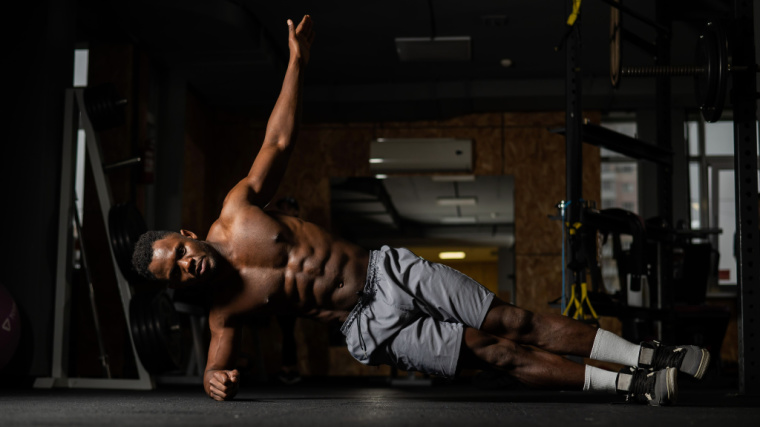 A shirtless person performs a side plank with their upper arm extended in the gym.