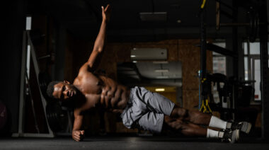 A shirtless person performs a side plank with their upper arm extended in the gym.