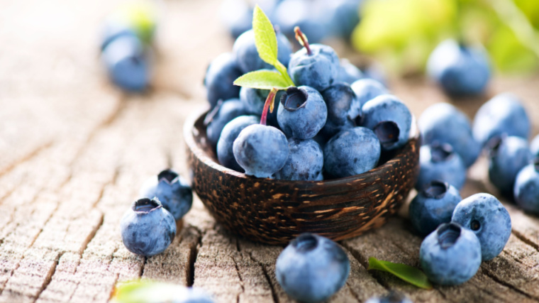 Blueberries sit scattered on a wooden table and in a small woven brown bowl.