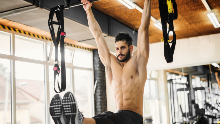 A shirtless person performs an L-sit pull-up in the gym.