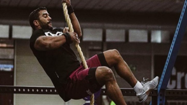 Kealan Henry wears a black t-shirt and maroon shorts while performing a rope climb.