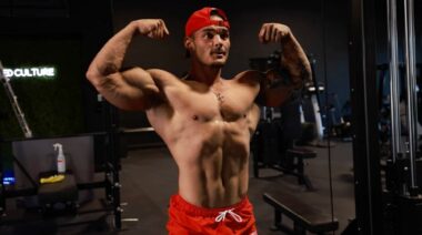 Shirtless bodybuilder in a red hat posing.