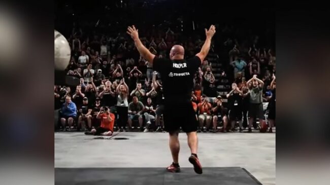 Person wearing a black shirt raises their hands in front of a crowd at a strongman competition.