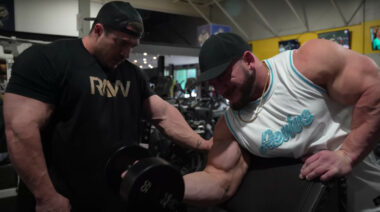 Two bodybuilders. One of which is curling a dumbbell in the gym.