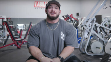 A bodybuilder in a gray shirt and black hat smiling at the camera.