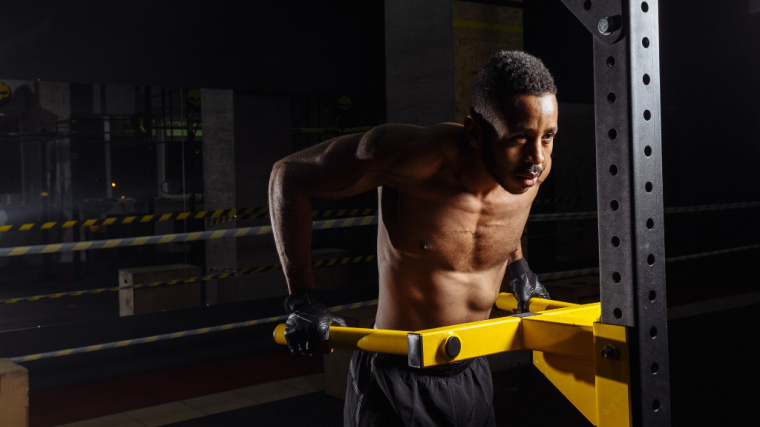 A shirtless person performs dips on a yellow dip bar.