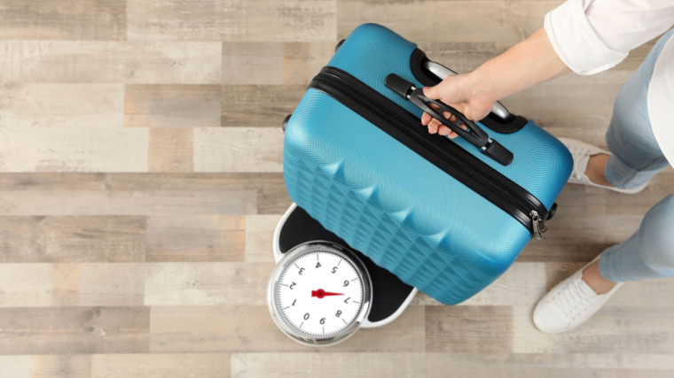 A close-up image shows a person holding a blue suitcase steady while they weight it on a home scale.