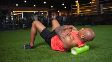 A person with a tattoo sleeve and an orange tank top crosses their arms over their chest while stretching their upper back over a neon green foam roller.