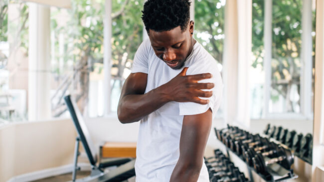 A person wearing a white t-shirt holds their shoulder in pain while standing in a gym.