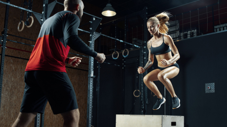 A person wearing a sports bra performs a box jump while their coach cheers them on.