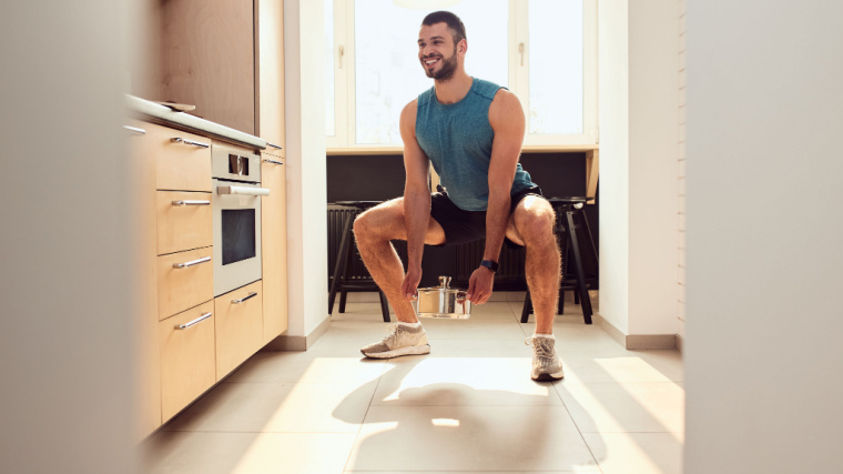 A person wearing a tank top and shorts performs a squat while holding a pasta pot in their kitchen.