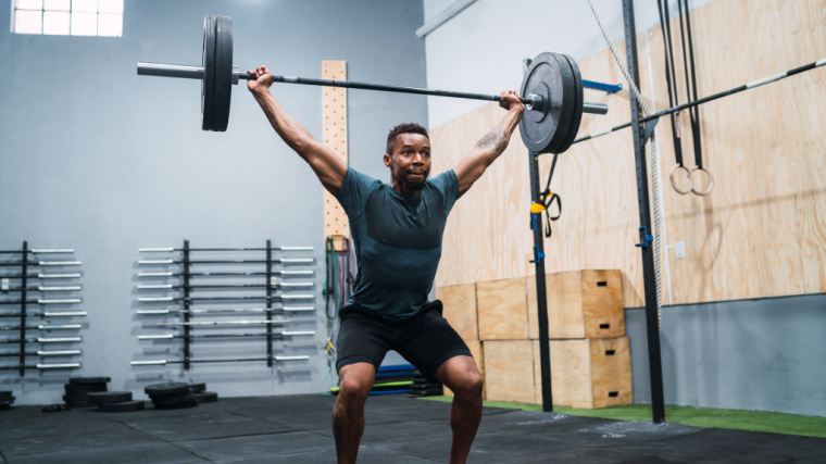A person wearing a blue t-shirt lifts a bar overhead with a snatch grip in a partial squat.