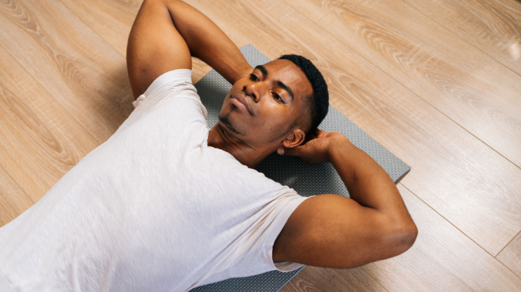 A person wearing a white t-shirt prepares to perform a crunch with their hands behind their ears.