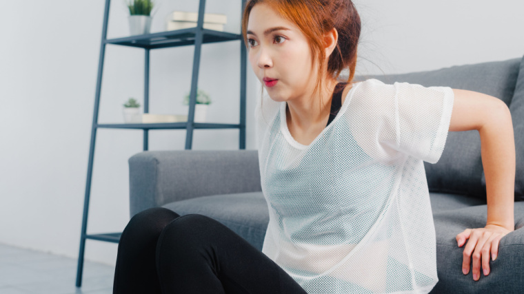 A person wearing a sheer white shirt and black sports bra performs a dip using their couch at home.