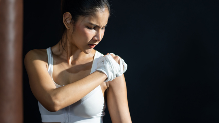 A person with white wraps for boxing loosely on their hands holds their hand to their shoulder in pain.