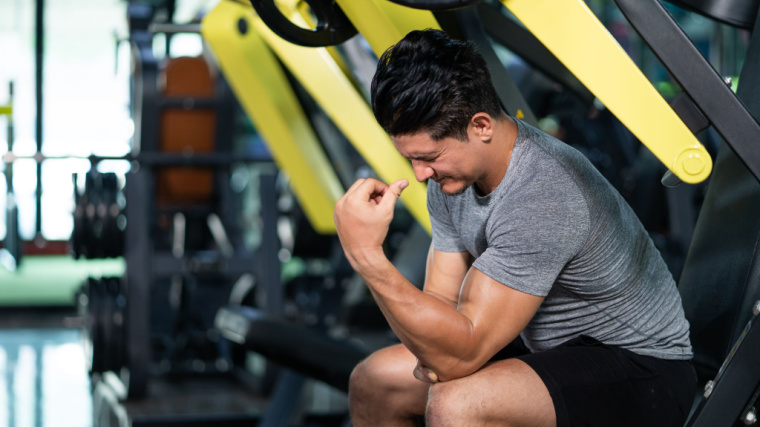 A person wearing a grey t-shirt grimaces in pain at their forearm and wrist while sitting on a bench in the gym.