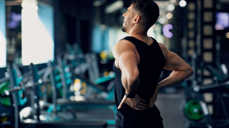 A person wearing a black tank top faces away from the camera with their hands on their low back, looking pained.