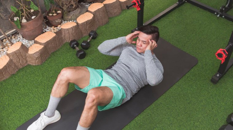 A person wearing a long-sleeved grey shirt and bright green shorts peforms a crunch on turf in a gym.