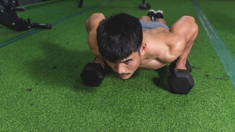 A person wearing a light blue muscle tank performs a push-up with dumbbells in their hands.