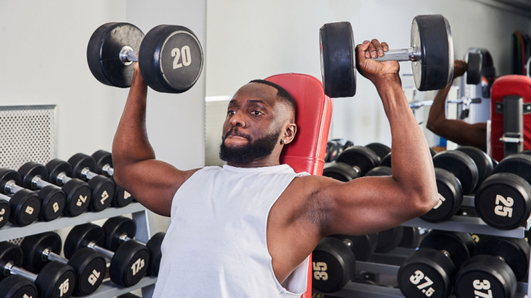 A person wears a white tank top and performs a seated shoulder press with 20-pound dumbbells.