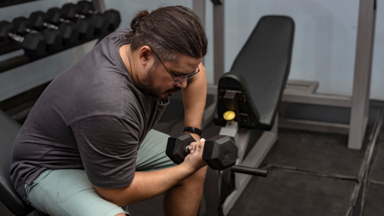 A person with facial hair keeps their hair in a bun while performing a concentration curl in the gym.