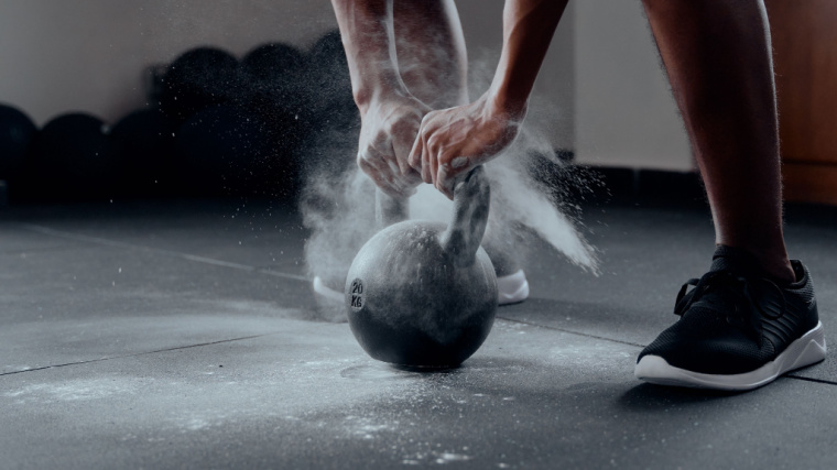 A close-up image shows a person grasping a single kettlebell with both hands with a lot of chalk floating around them.