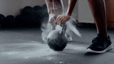 A close-up image shows a person grasping a single kettlebell with both hands with a lot of chalk floating around them.