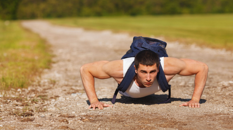 A person wearing a blue backpack performs push-ups on a dirt track.
