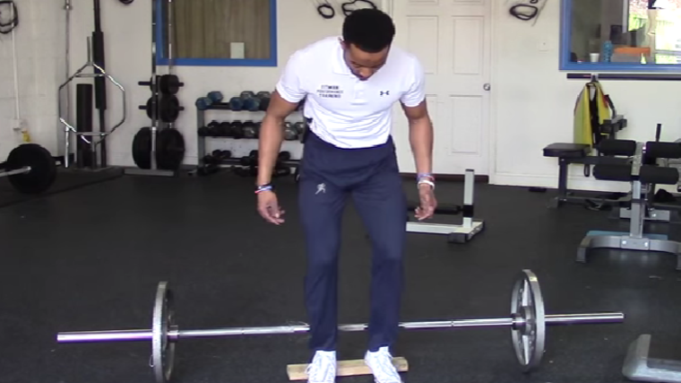 A person wearing a white polo steps over a barbell and prepares to perform a hack squat.