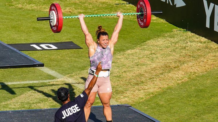 CrossFitter Kara Saunders pressing a green barbell loaded with red plates overhead on a turf field.