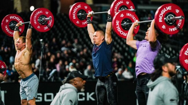 CrossFit athletes pressing loaded barbells overhead with red plates on each side on a turf field.