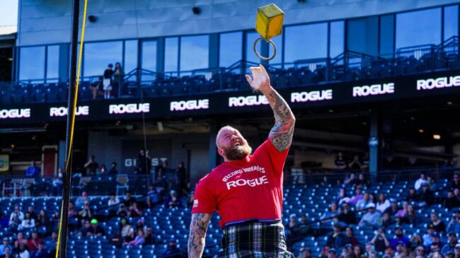 Strongman Hafthor Bjornsson, wearing a kilt and tossing a weight over a tall, yellow bar