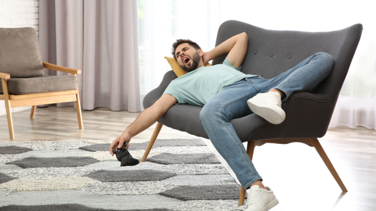 A male person lying down on couch yawning holding a video game controller.