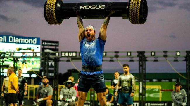 CrossFitter Tim Paulson pressing a strongman log overhead while wearing a blue shirt, black shorts, and in front of other athletes at the 2022 Rogue Invitational.