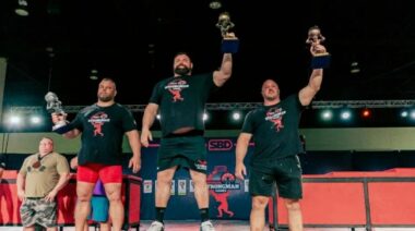Strongman stand atop a podium holding trophies overhead.