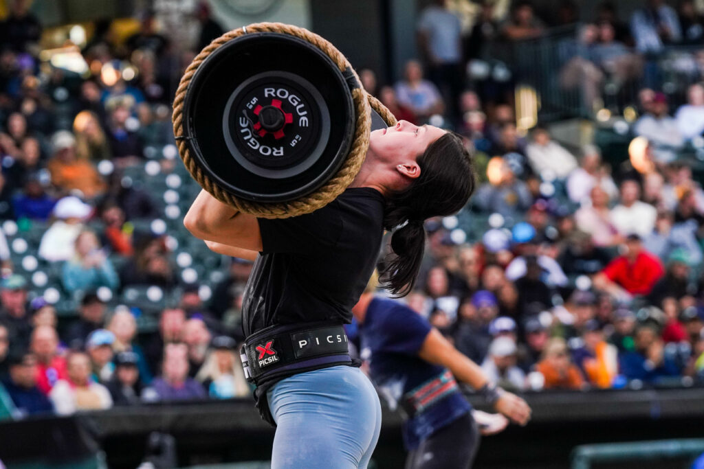 Emma Lawson, wearing a black t-shirt and blue leggings, holds a strongman log across her upper chest.