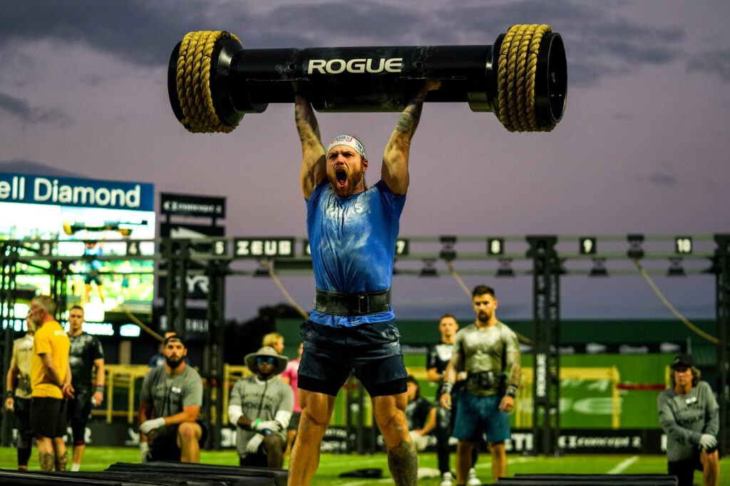 CrossFitter Tim Paulson pressing a strongman log overhead while wearing a blue shirt, black shorts, and in front of other athletes at the 2022 Rogue Invitational. 