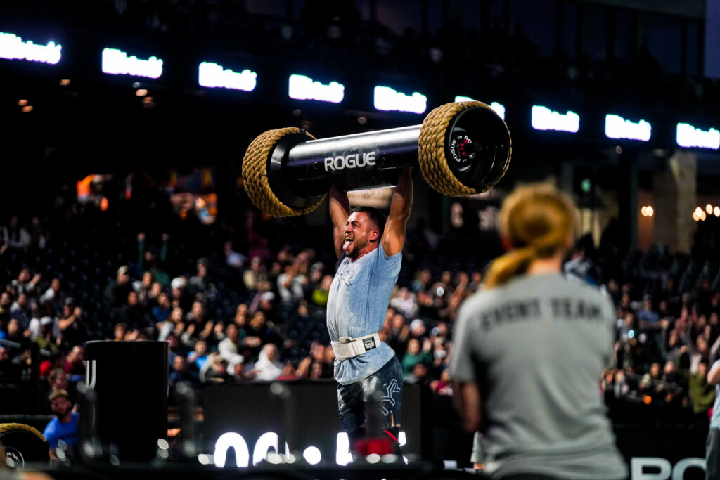 Ricky Garard dons a blue shirt and black workout shorts as he presses a heavy strongman log over his head.