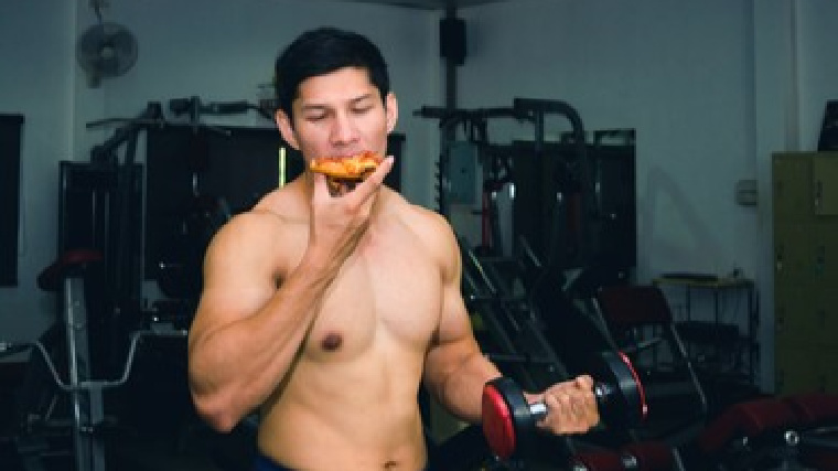 A shirtless person eats a slice of pizza while holding a dumbbell.