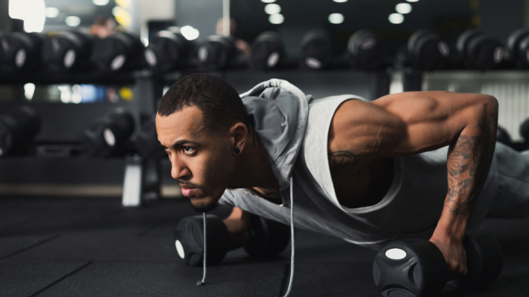 A person wearing a sleeveless hoodie performs a push-up while holding dumbbells.