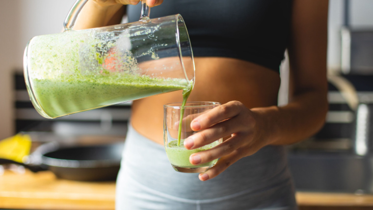 A person pours a green smoothie from a pitcher into a glass.